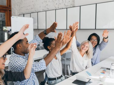 Adorable girl with short blonde hairstyle slapping hands with african female colleague. Indoor photo of joyful coworkers celebrating beginning of vacation at workplace.